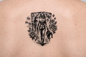 A black-and-grey rib tattoo showing Harley walking forward with her back turned to a torn Joker card behind her. One broken chain falls from her wrist, while her jester hood hangs loosely from her hand—showing that she is leaving the old version of herself behind. The words “Ha Ha Ha” fade near the Joker card, while diamonds and roses flow upward alongside Harley. tattoo idea