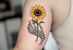 A skeleton hand holding a dying yellow sunflower, "memento mori" in thin, winding black cursive along the stem. tattoo idea