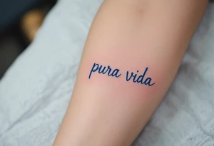 Simple cursive “Pura Vida” inked in navy blue with a small lavender sprig underlining the text. tattoo idea
