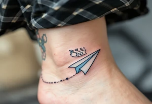 A cartoon-style paper airplane in soft grey and baby blue, trailing a banner with the birthdate 08. 12. 2022. tattoo idea