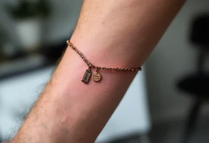 A dainty chain bracelet tattoo with hanging charm tags engraved with "Kristian – 15.8.2018 v 21:21" in metallic bronze ink tattoo idea