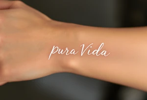 “Pura Vida” in white ink across a tan skin tone, with subtle shadowing for a sun-kissed effect. tattoo idea