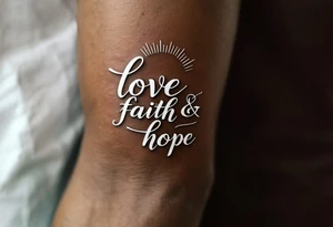 Tattoo in white ink on dark skin, featuring delicate serif lettering and a silver-lined halo above the word "love faith hope. tattoo idea
