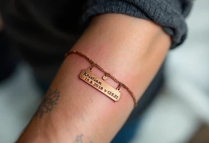 A dainty chain bracelet tattoo with hanging charm tags engraved with "Kristian – 15.8.2018 v 21:21" in metallic bronze ink tattoo idea
