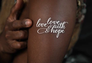 Tattoo in white ink on dark skin, featuring delicate serif lettering and a silver-lined halo above the word "love faith hope. tattoo idea