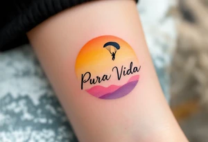 Cursive “Pura Vida” trailing behind a paragliding silhouette against a sunset gradient of yellow, orange, and magenta. tattoo idea