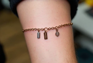 A dainty chain bracelet tattoo with hanging charm tags engraved with "Kristian – 15.8.2018 v 21:21" in metallic bronze ink tattoo idea