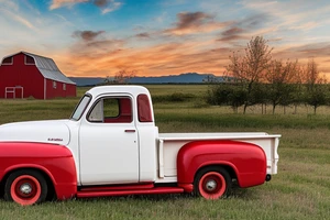 A rustic red and white '53 5-window GMC long bed pickup parked at an angle in front of a red barn with the setting sun in the background. The setting sun is shaped like a sunflower. tattoo idea